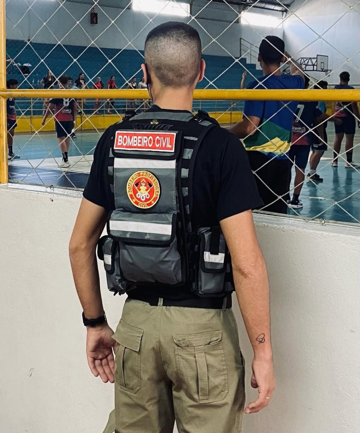 Bombeiro civil de costas, com colete, acompanhando uma atividade esportiva em um ginásio