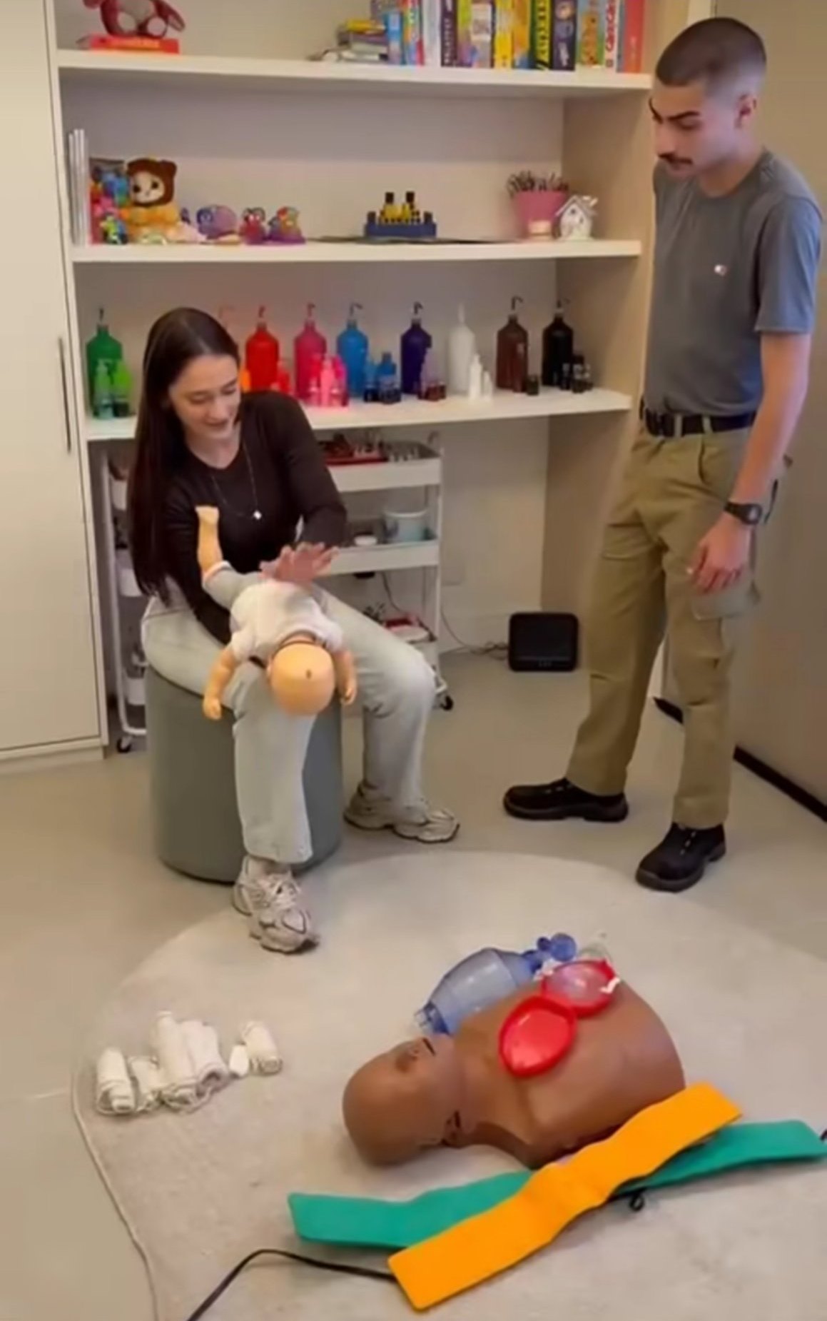 Aluna praticando com manequim de bebê enquanto Fernando acompanha, com manequim adulto e materiais no chão